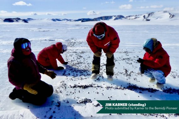 Four people wearing red arctic gear look at an ice-and snow-covered field. Three of the men are kneeling and one is bending over at the waist looking at the ground.