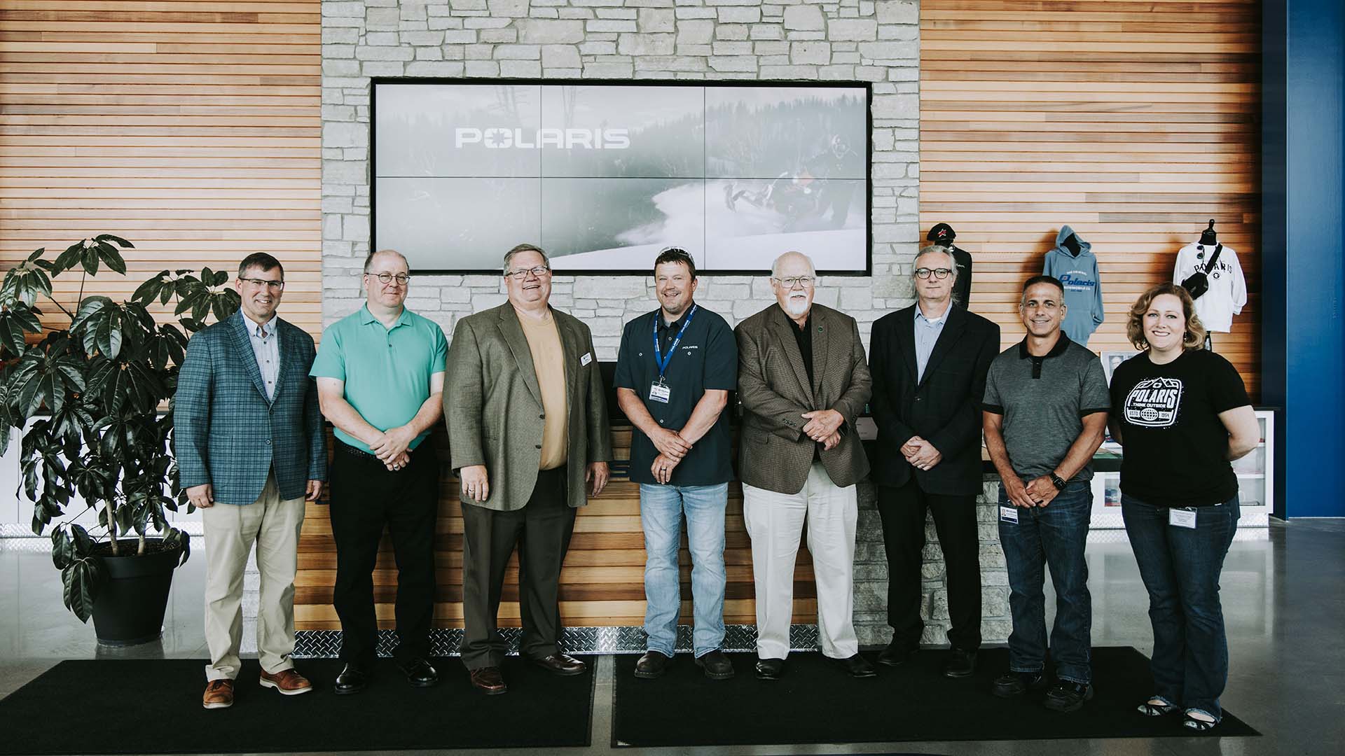 A group of eight people pose in front of a rock and wood wall with a bank of TVs displaying the Polaris logo.