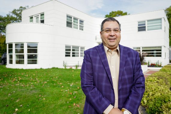 A man with glasses wearing a collared shirt and a plaid purple sportcoat stands in front of the David Park House at BSU. It is a white house with a flat roof.