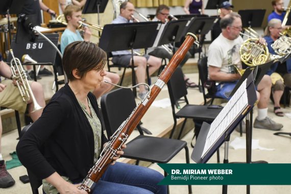 Miriam Webber plays an oboe; she is wearing a black cardigan and jeans in a room full of other band musicians