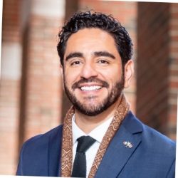 A photo of a smiling man with a beard. He is wearing a blue suit with a dark blue tie and he has a brownish-gold scarf.