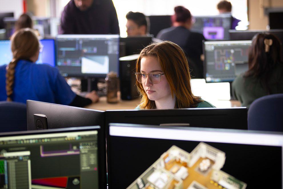 A student with reddish brown shoulder-length hair and glasses works at a station in a computer lab.