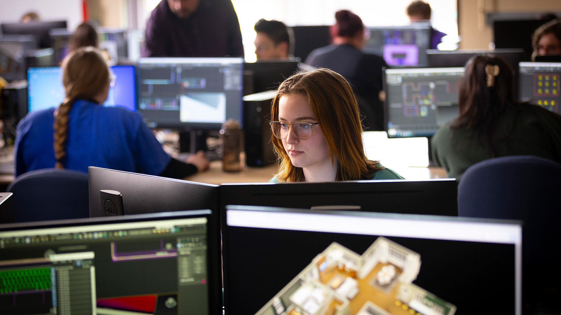 A student with reddish brown shoulder-length hair and glasses works at a station in a computer lab.